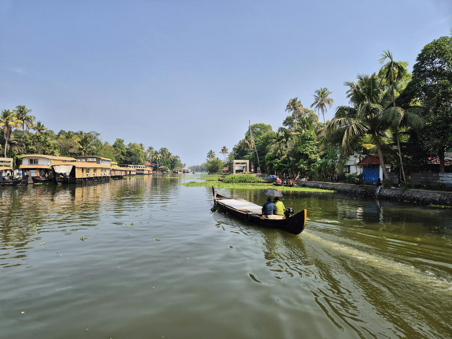 A serene view of a traditional boat navigating the lush Kerala backwaters with houseboats and palm trees in the background.