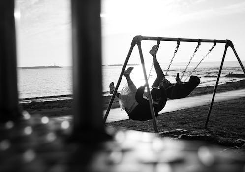 A couple enjoys swinging together by the seaside, capturing joyful moments in black and white.