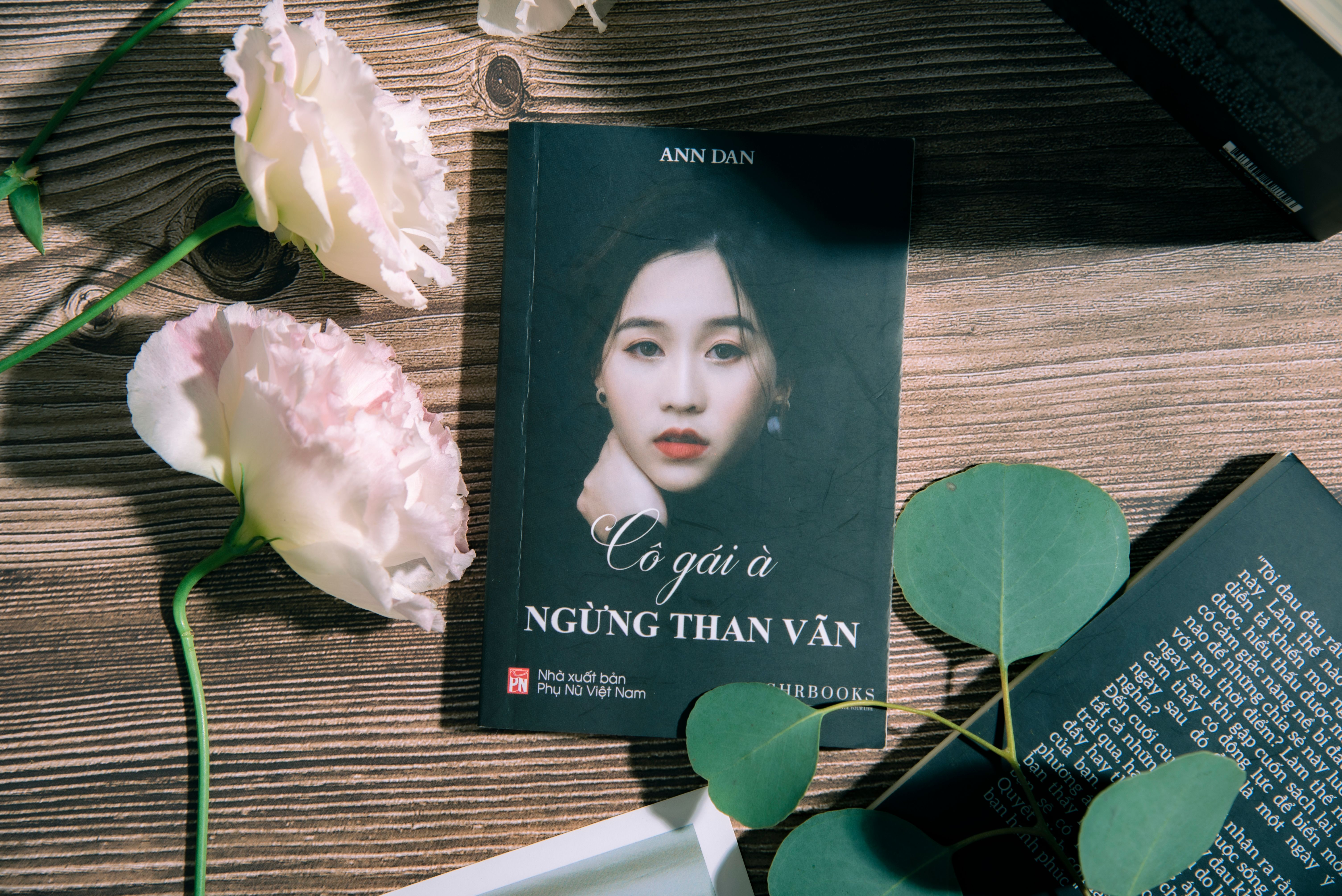 A book titled 'Cô gái à' lays next to elegant flowers on a wooden table.