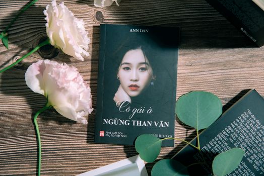 A book titled 'Cô gái à' lays next to elegant flowers on a wooden table.