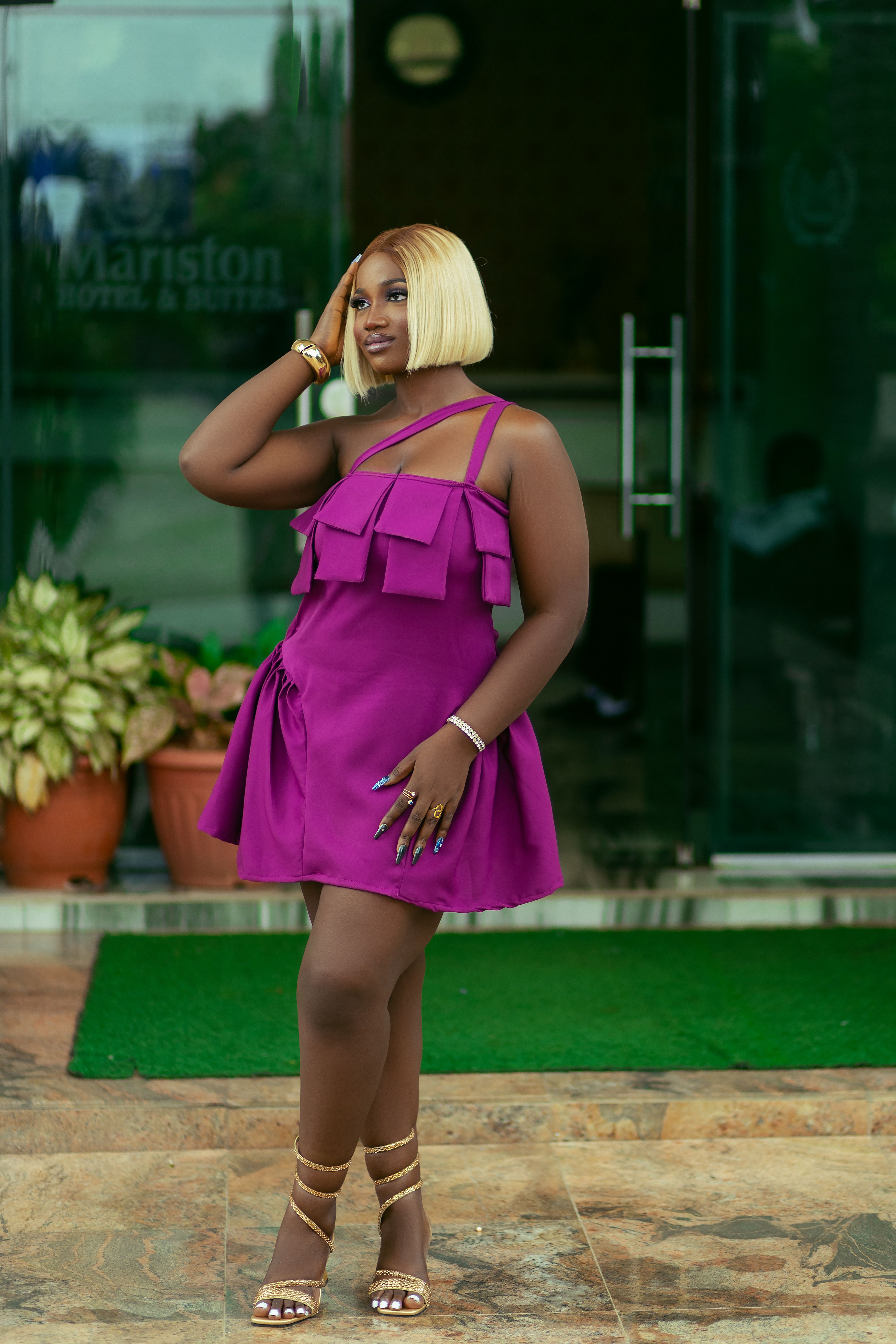 Elegant woman poses in purple dress at hotel entrance, showcasing fashion style.