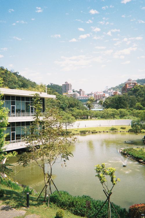 Free Urban park featuring a serene lake, modern buildings, and lush greenery under a clear sky. Stock Photo