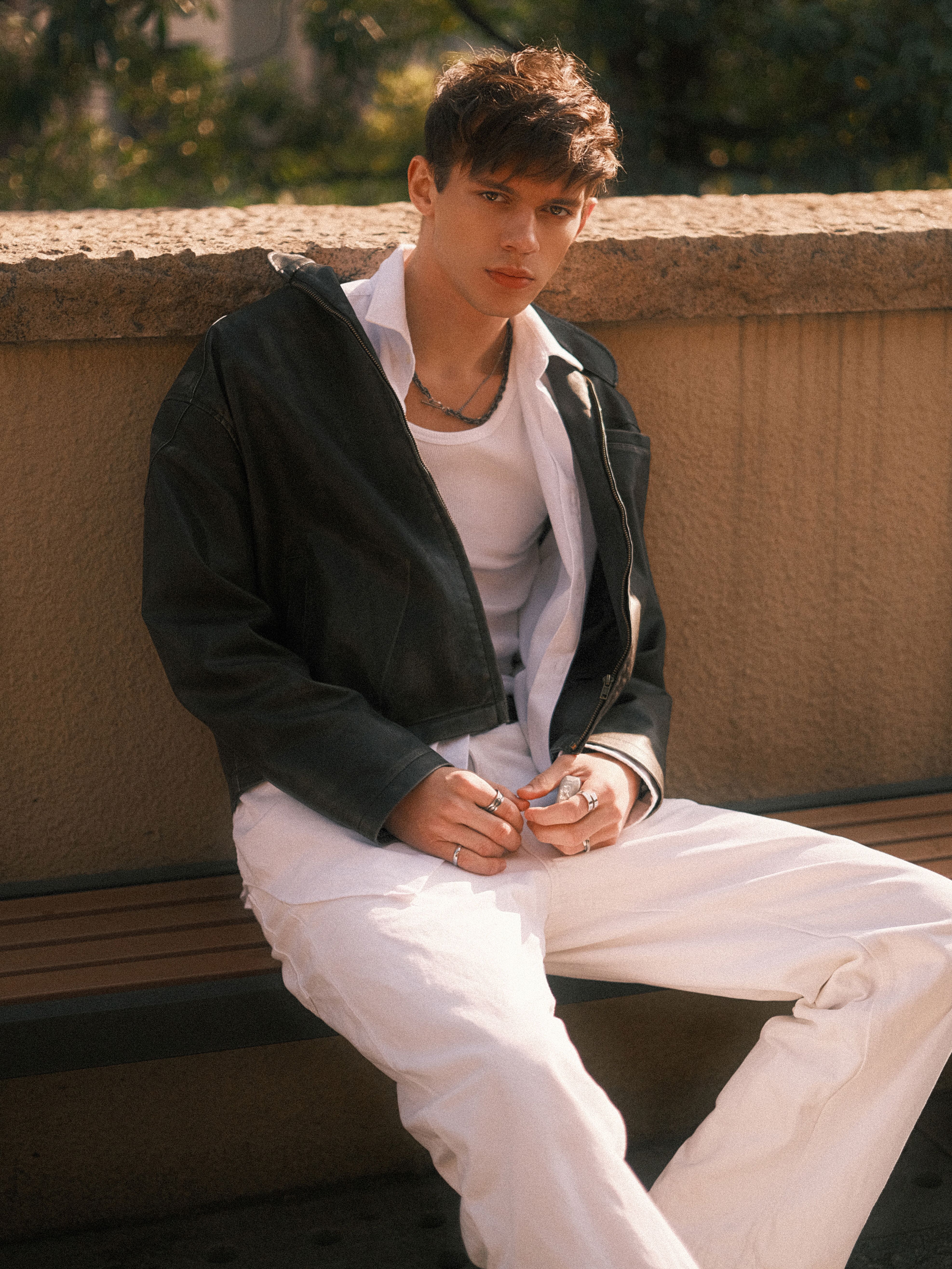 Stylish young man sitting outdoors in casual attire, expressing a relaxed mood.