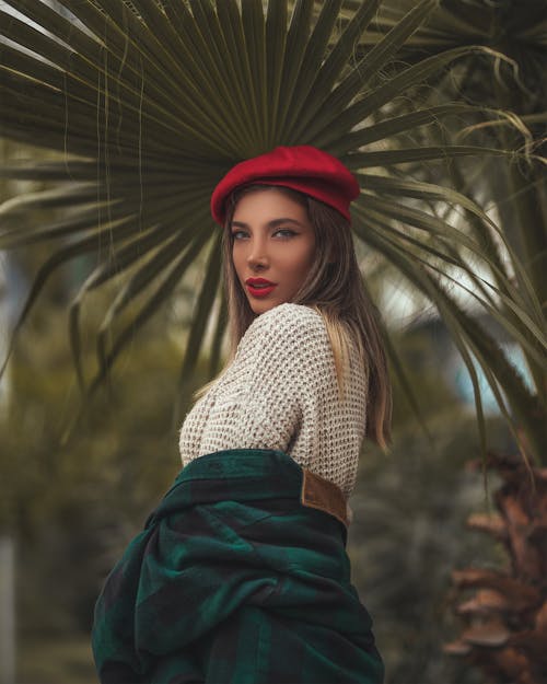 Selective Focus Photo of Woman in Red Hat Posing Under Palm Tree Leaf