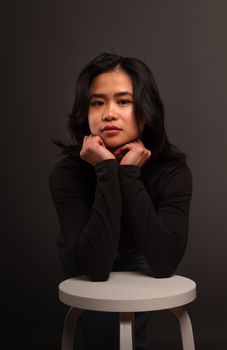 A contemplative young woman poses indoors, leaning on a stool in a dark setting.