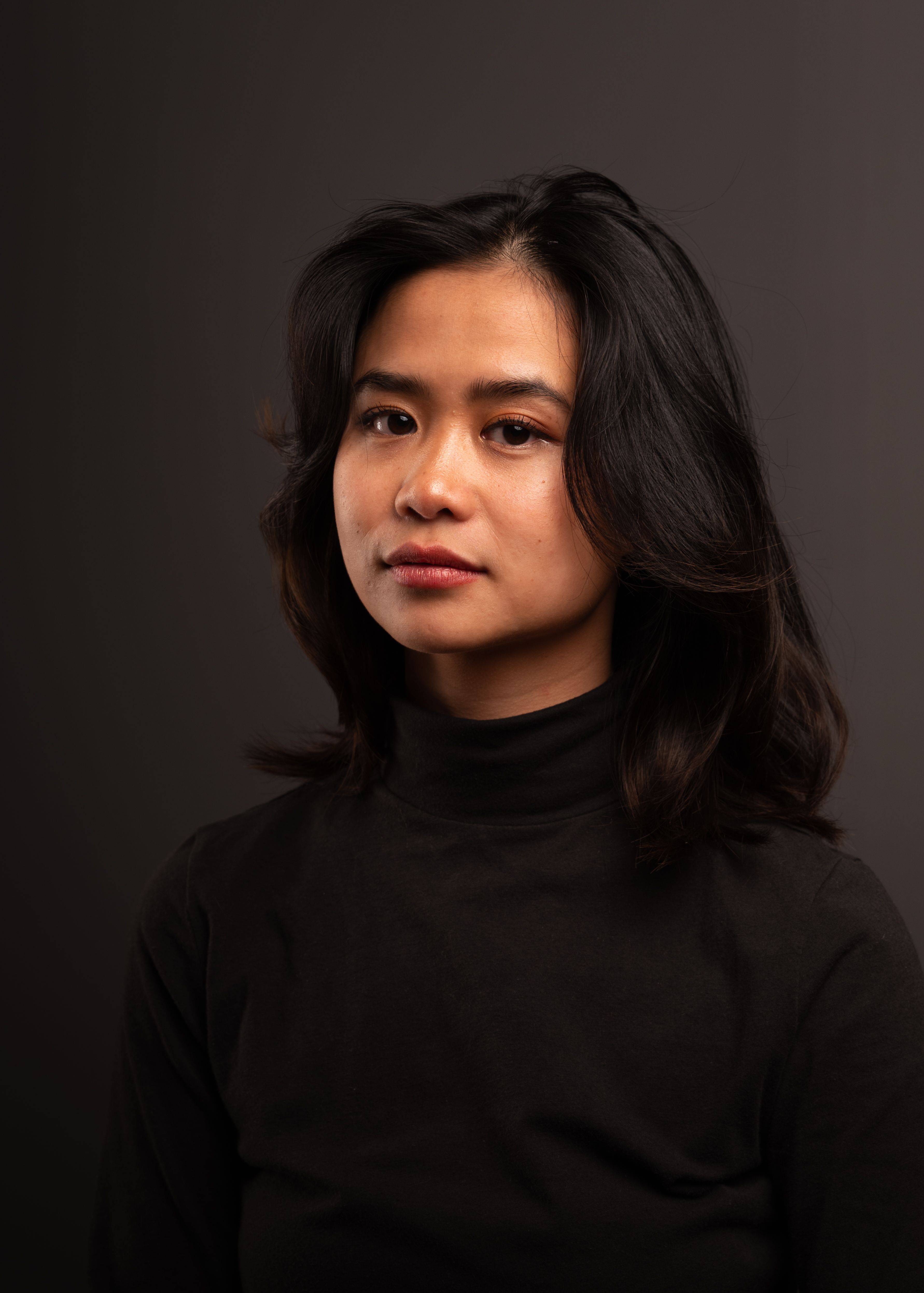 A close-up portrait of a woman with dark hair wearing a black turtleneck against a dark background.