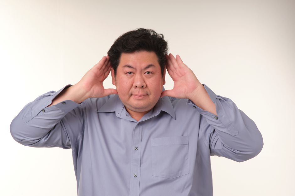 ニュートラルな背景を背景に、青いシャツを着て、耳に手を当ててジェスチャーをしている男性のスタジオポートレート。