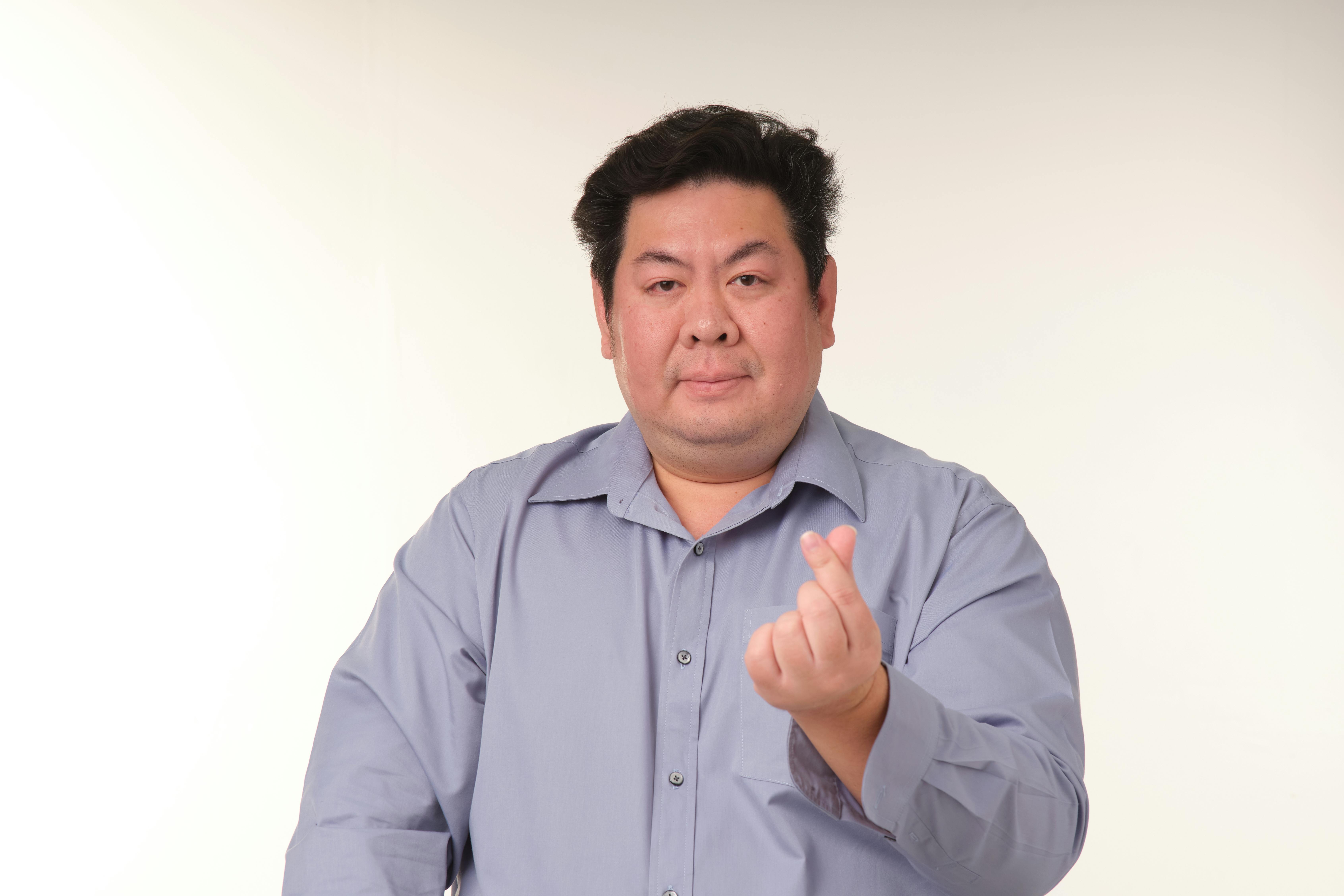 Kostenlos Mann mittleren Alters in blauem Hemd macht vor schlichtem Hintergrund eine Herzgeste mit dem Finger. Stock-Foto