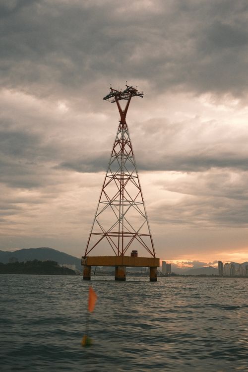 Free A tall electrical tower stands above a body of water during sunset with moody skies. Stock Photo