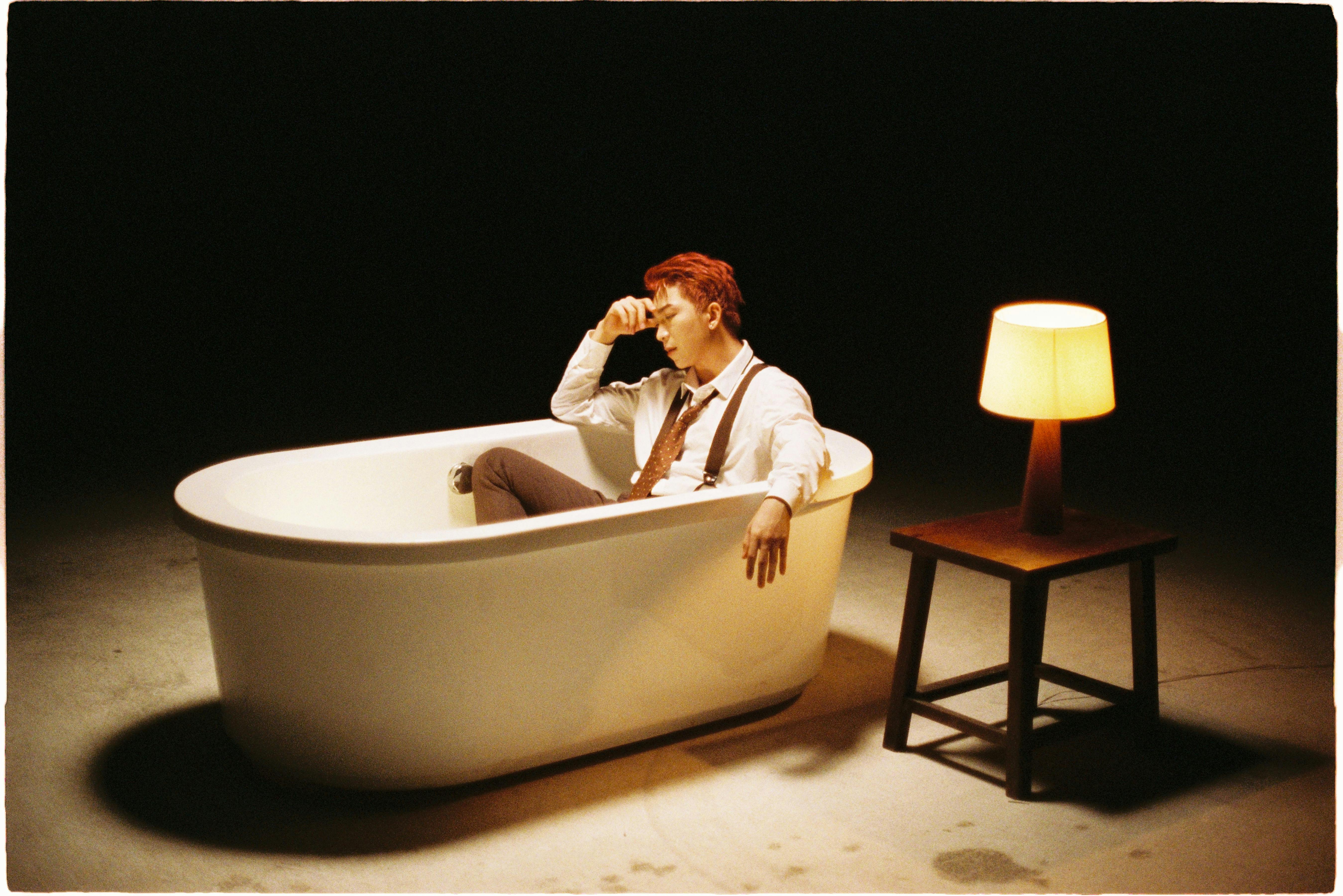 Dramatic scene of man sitting in a bathtub next to a lamp, evoking introspection.