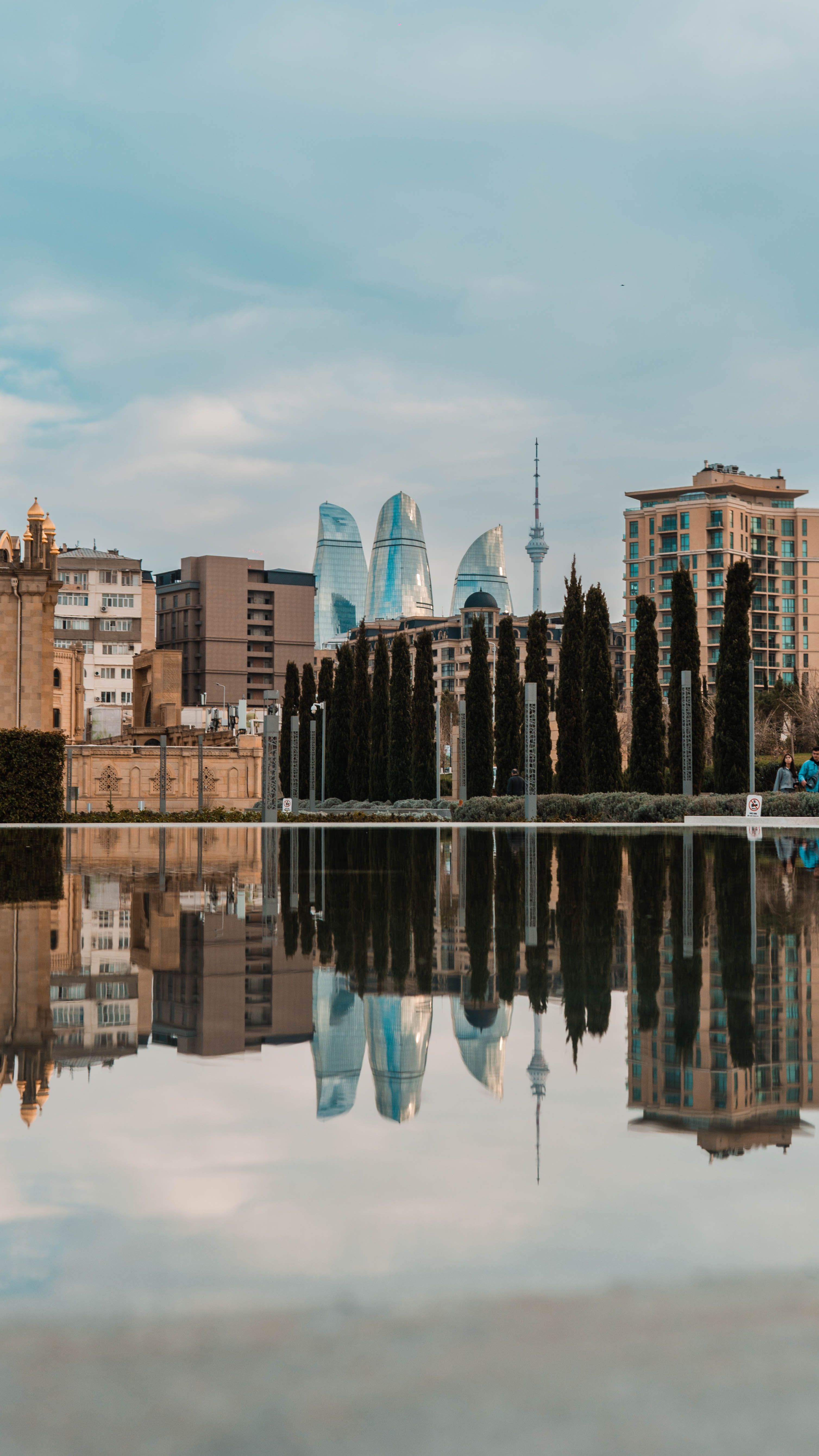 Scenic view of Baku's Flame Towers reflecting in water with skyline and trees.