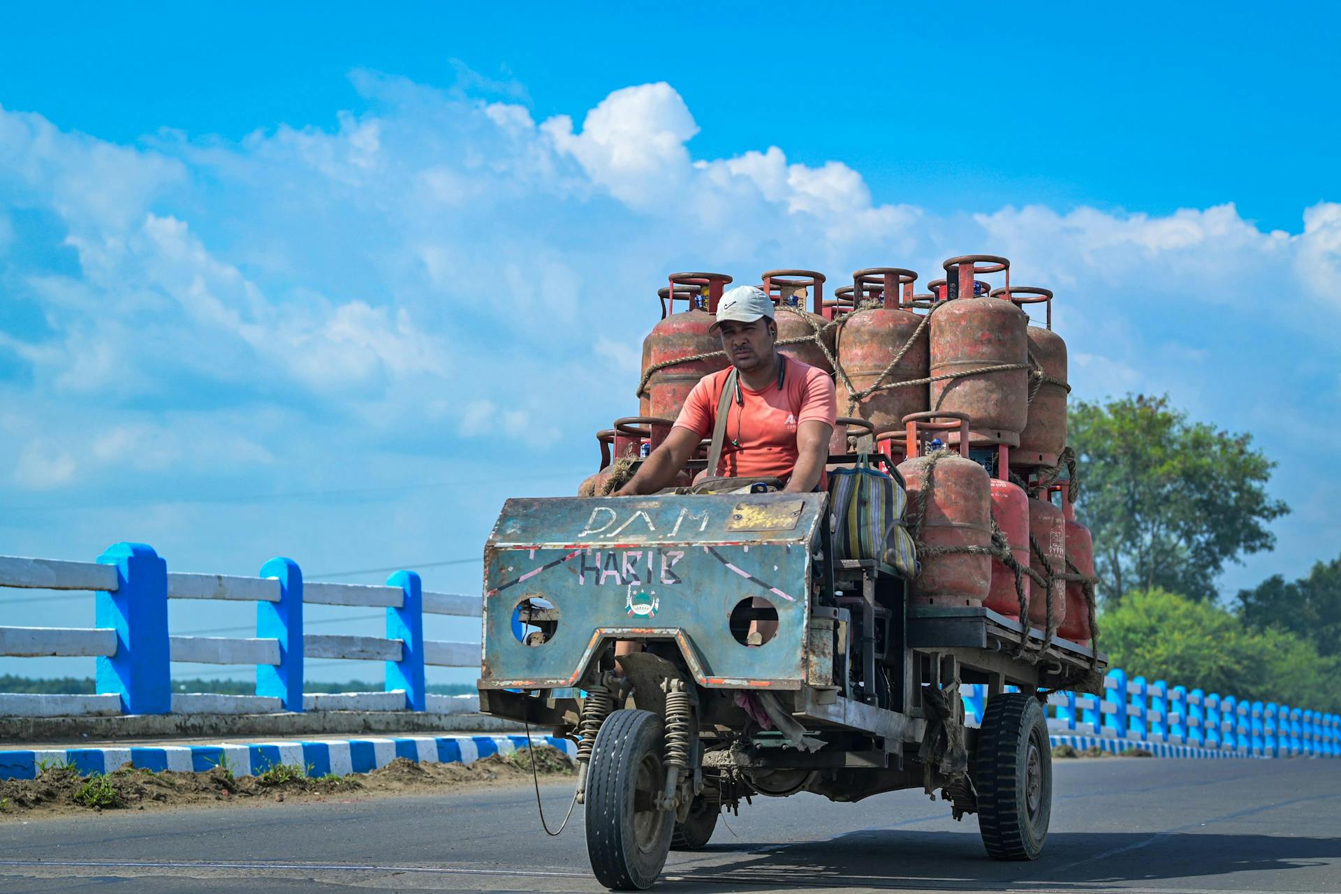 A man on a tricycle transporting gas cylinders on a sunny day over a bridge.