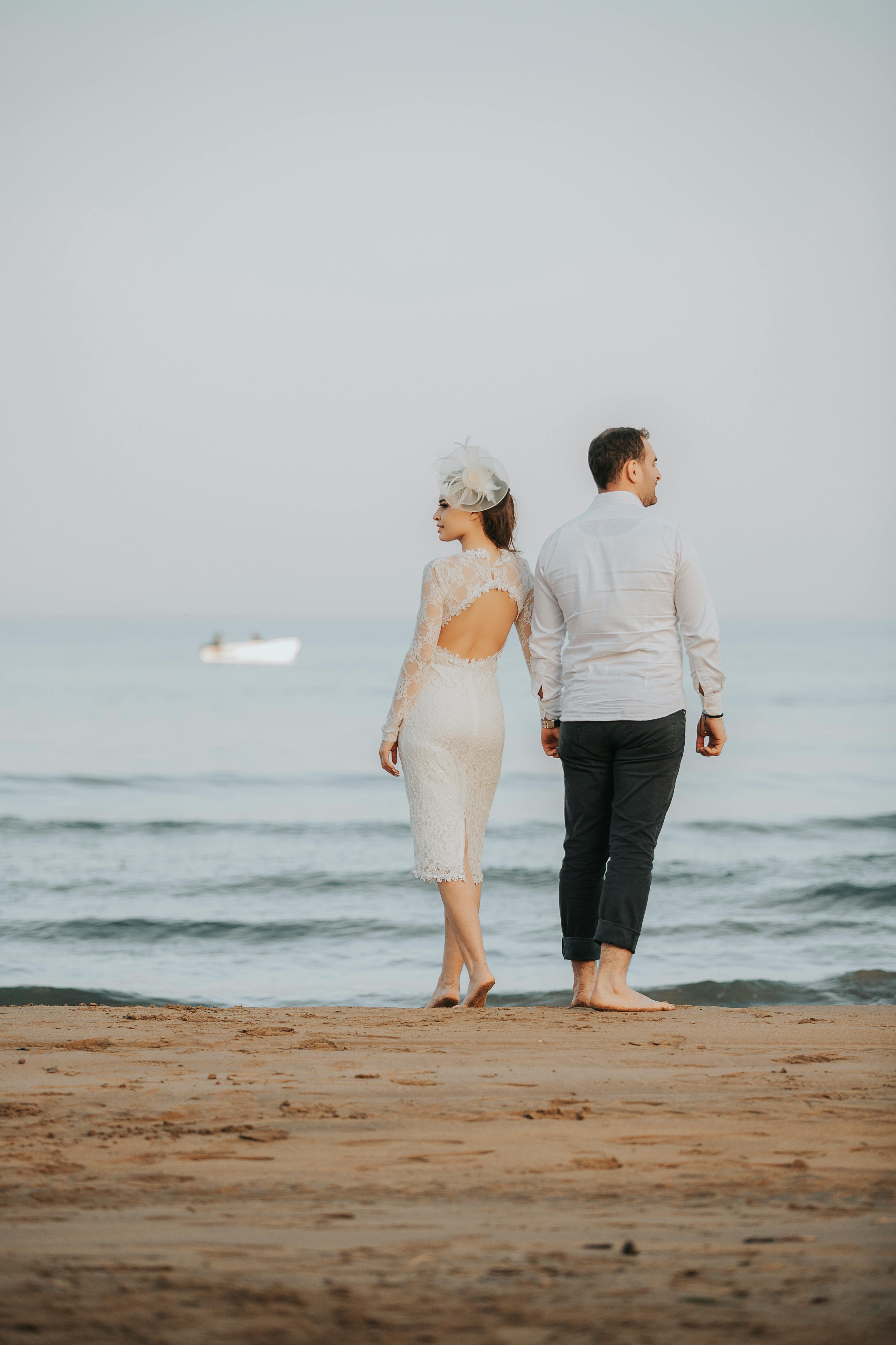 A couple dressed elegantly stands back to back on a beach, gazing at the calm sea during a serene day.