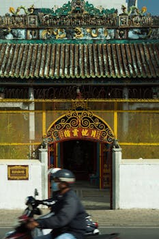 A motorcyclist drives by a traditional Asian temple gate with intricate designs.