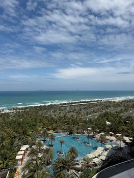 Sky-high view of a tropical resort with a pool, palm trees, and ocean waves under a blue sky.