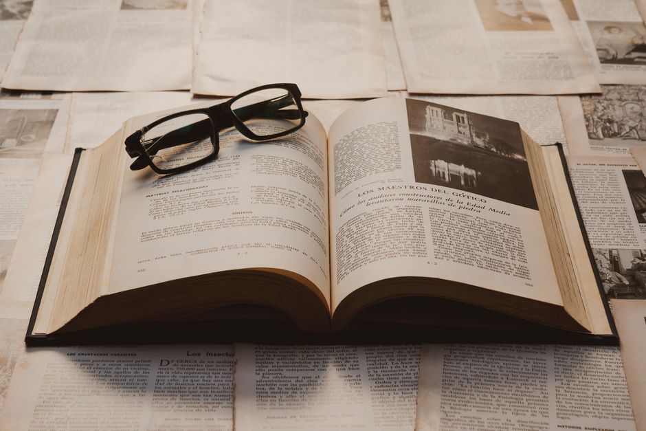 Un libro antiguo abierto sobre una mesa, rodeado de papeles viejos y con vasos apoyados.