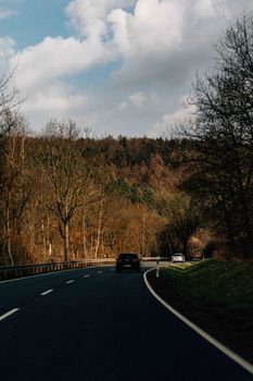 A car travels on a winding road through a picturesque autumn landscape with bare trees and cloudy skies.
