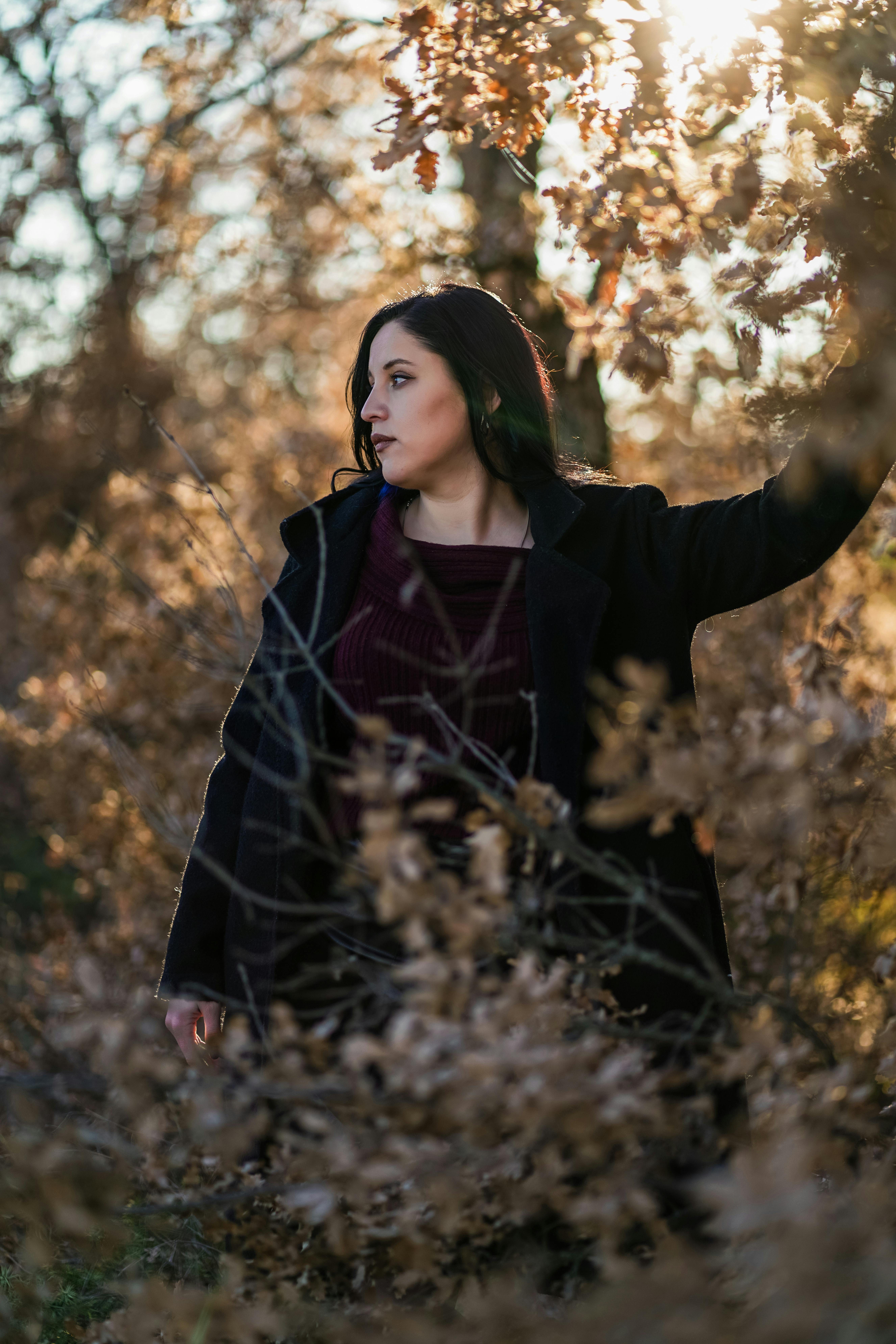 Woman in black coat amidst autumn leaves with warm sunlight filtering through.