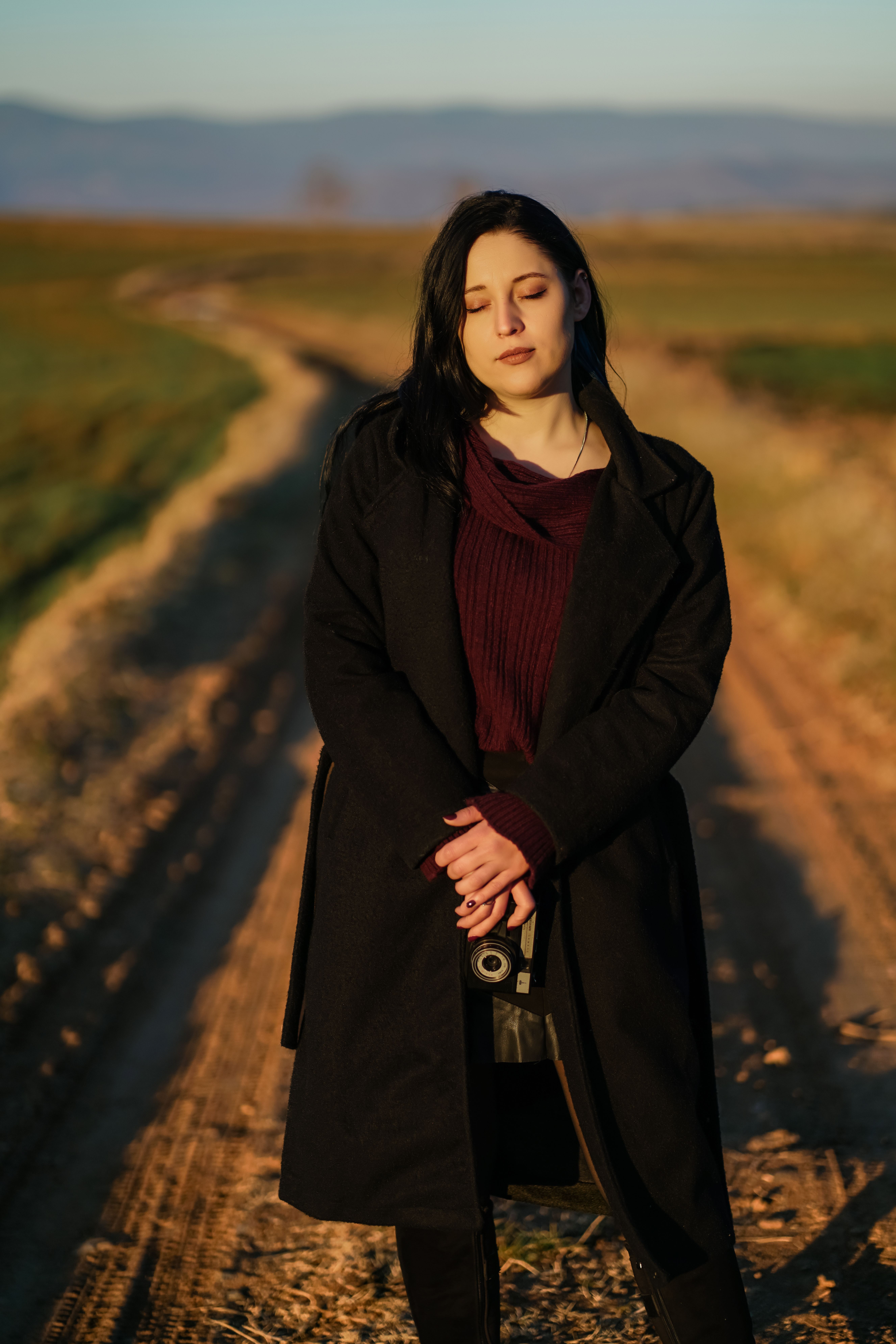 A woman in a dark coat stands contemplatively on a country road at sunset.