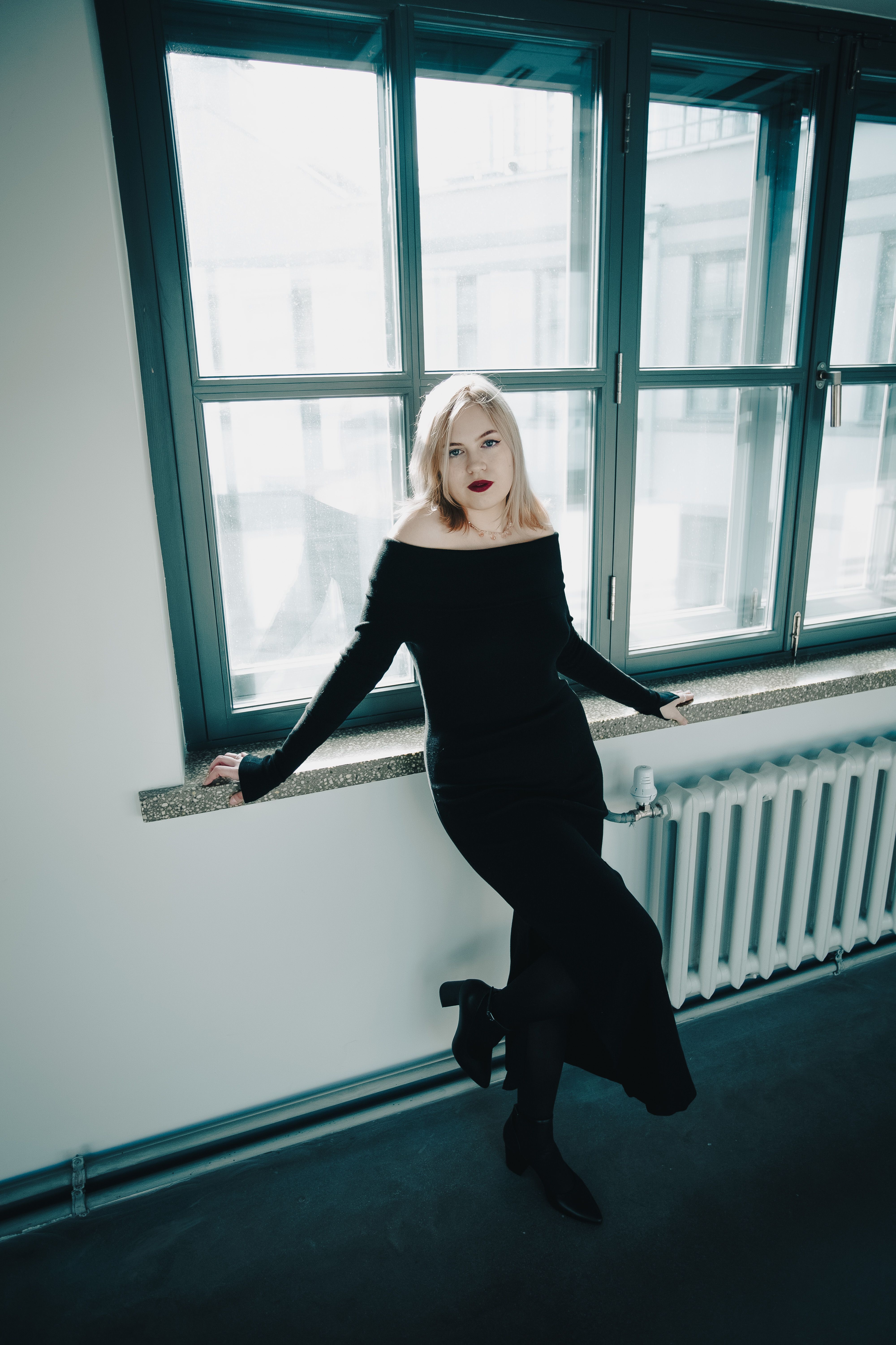 Stylish woman in black dress sits by a large window in a bright room.