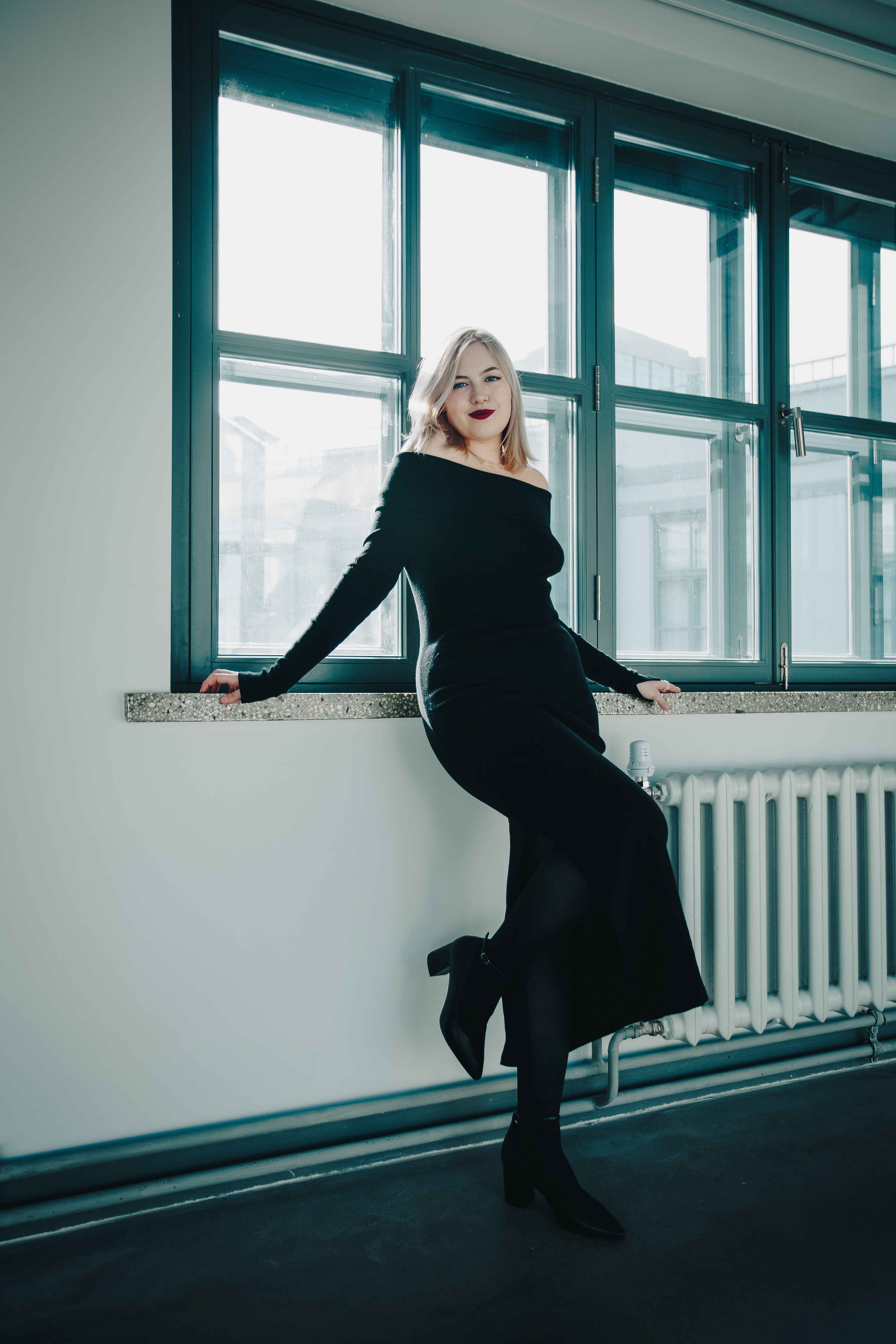 Stylish woman in a black dress poses by a large window in an indoor setting.