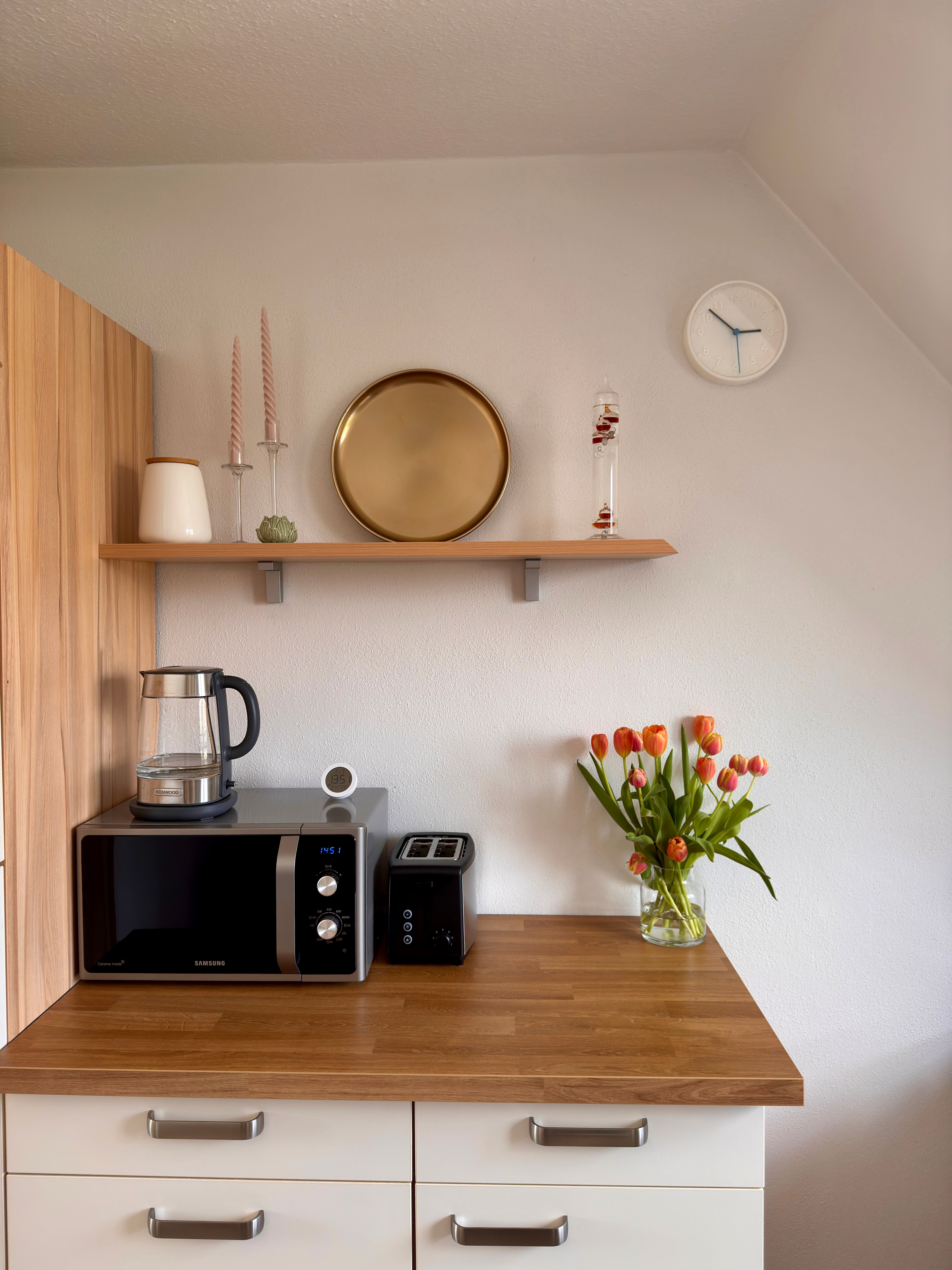 Bright kitchen corner with a small black two-slice toaster beside a microwave on a wooden worktop, tulips in a vase and a shelf above
