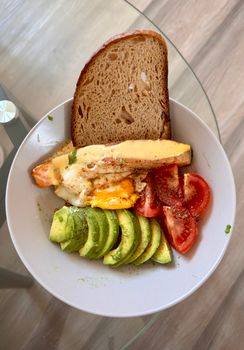 Top view of a nutritious breakfast plate with avocado, cheese, and bread on a glass table.