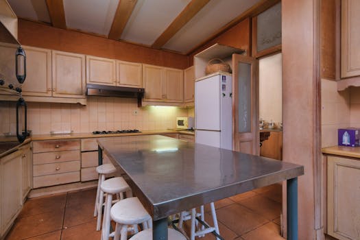 A rustic kitchen featuring wooden cabinets, stainless steel table, and stools, creating a cozy ambiance.