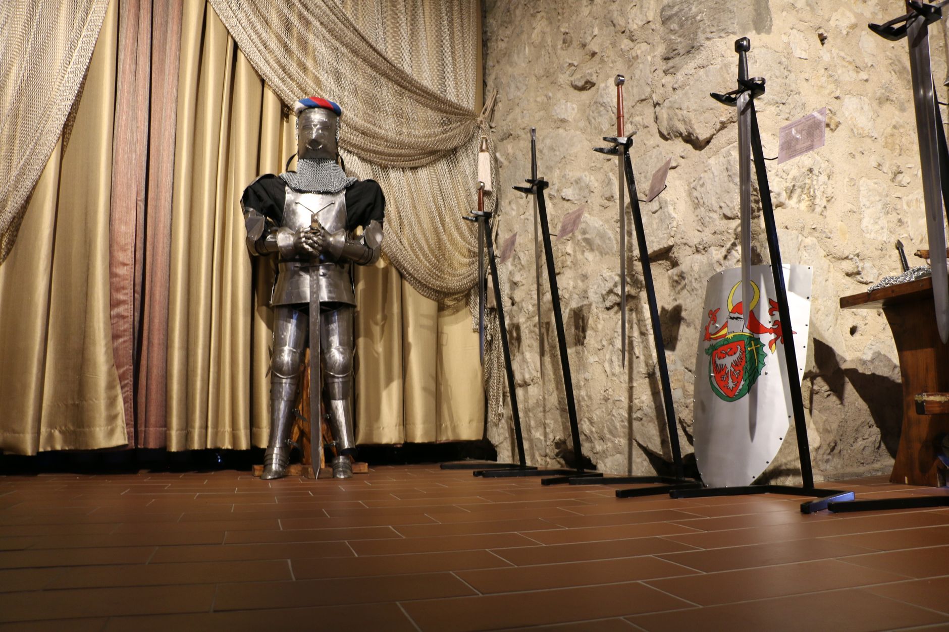 A knight's armor and swords displayed in a historic room with stone walls.
