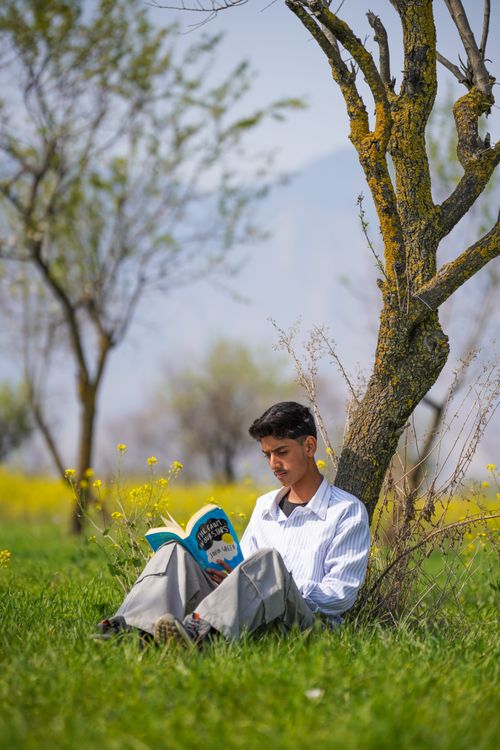 Free A young man reads a book while sitting under a tree in a lush green field during springtime. Stock Photo
