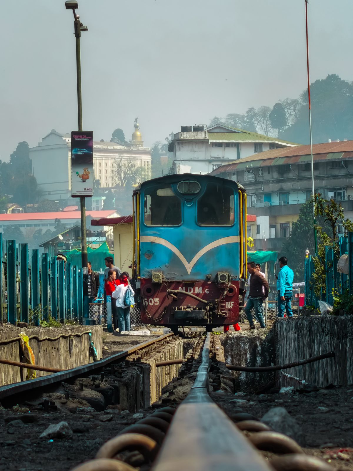 A blue narrow gauge locomotive waiting at Darjeeling station as travellers cross the track beside it