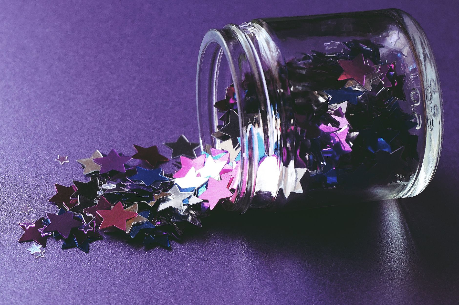 Close-up of colorful star-shaped confetti spilling from glass jar on purple surface. Ideal for creative and festive themes.
