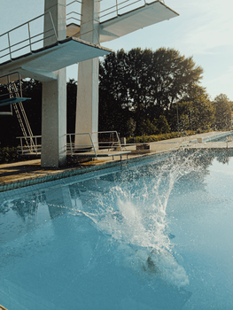 A high dive splash at an outdoor swimming pool under sunny skies.