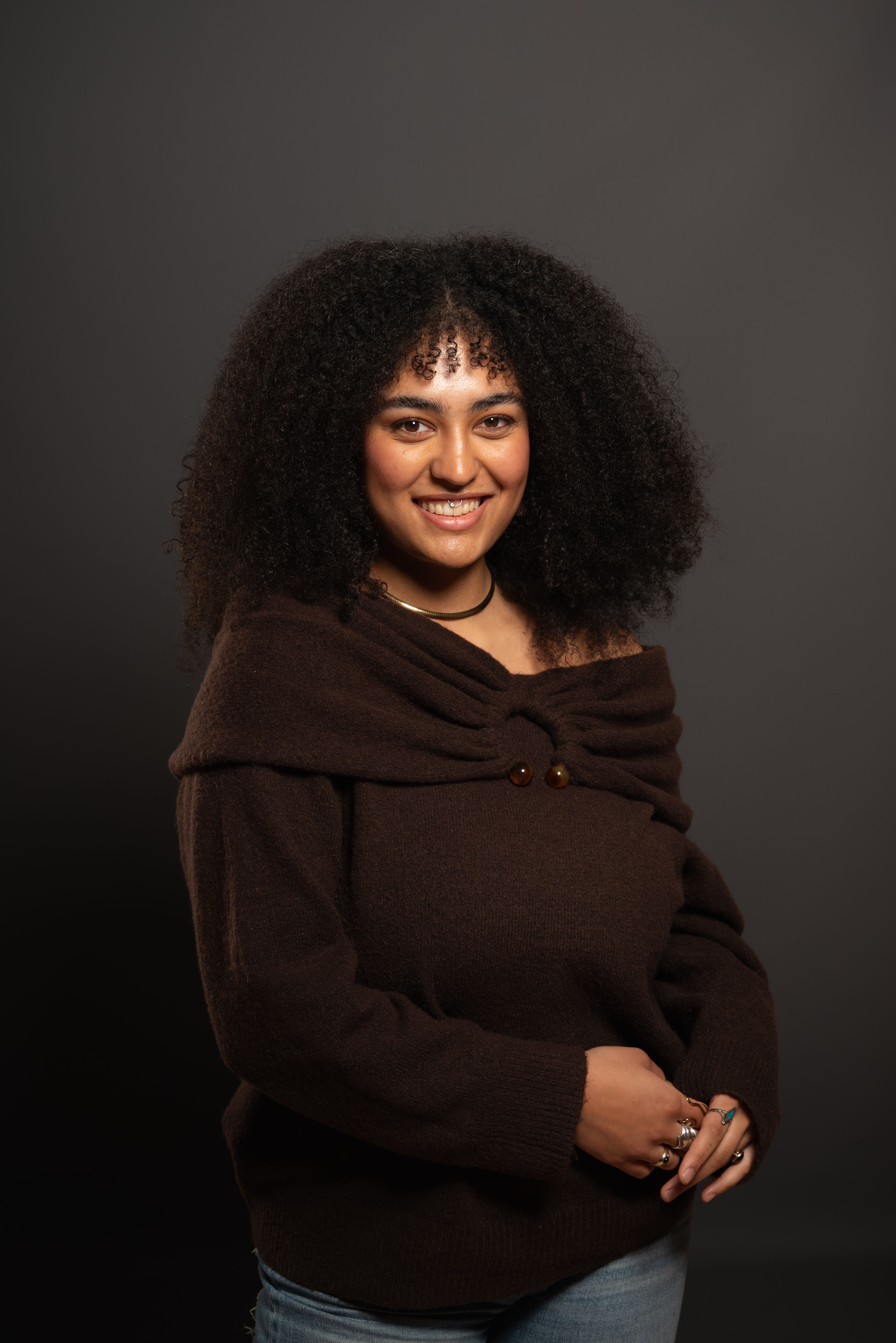 Portrait of a smiling woman with curly hair wearing a brown sweater against a gray background.