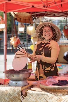 Cheerful woman in traditional attire cooking rice paper at a vibrant outdoor market stall.