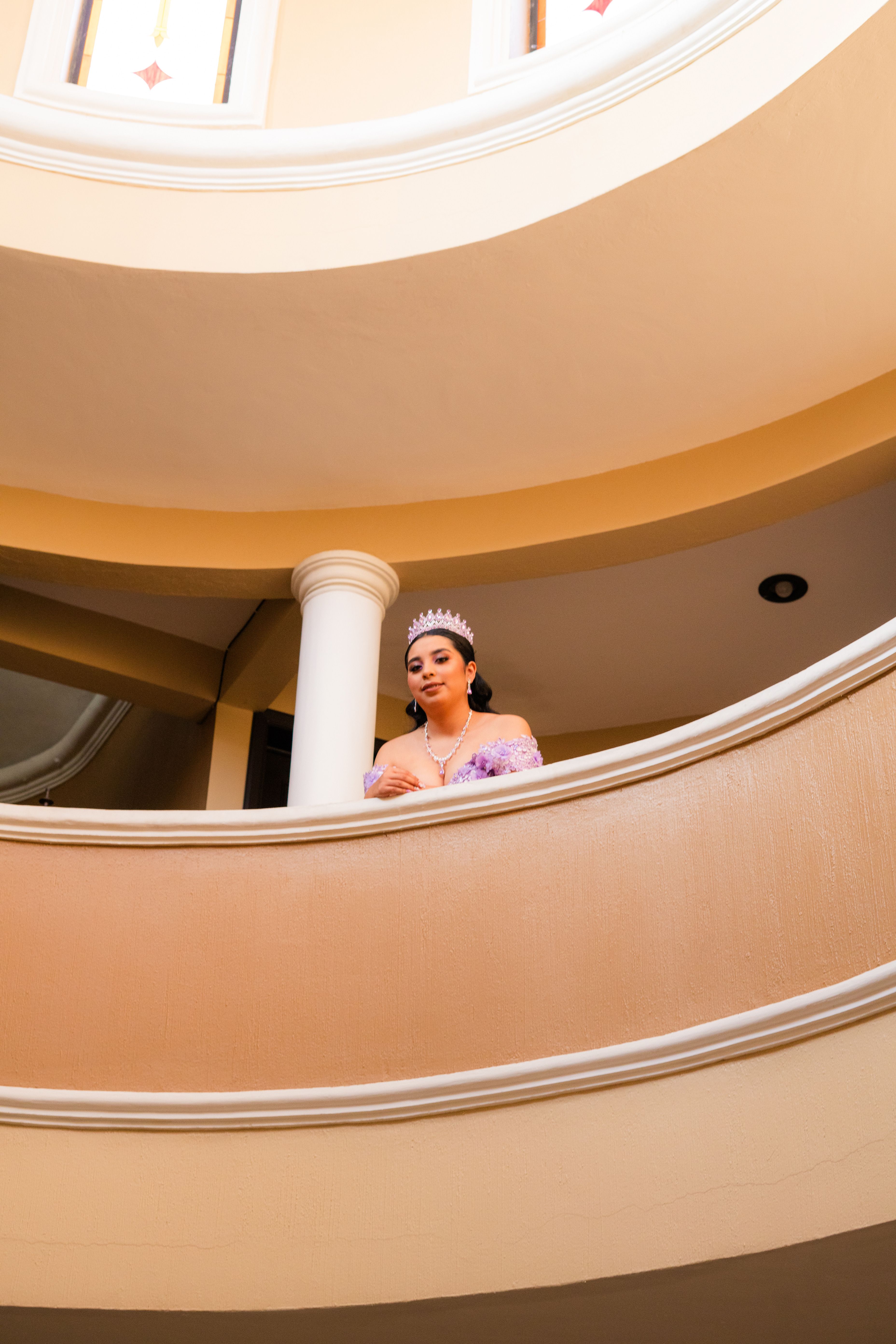 Elegant woman in formal dress and tiara standing on a curved indoor balcony.
