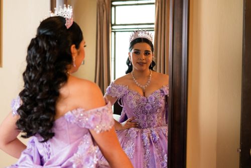 Free Young woman in a lilac Quinceañera dress with tiara looking into a mirror indoors. Stock Photo