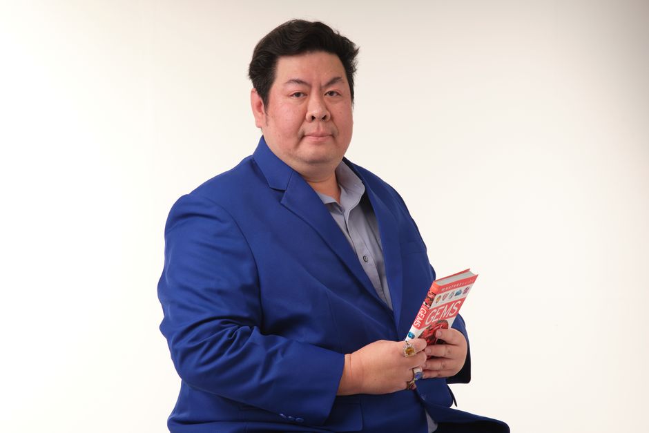 Adult man in blue suit holding a book about gems, seated indoors against a neutral background.