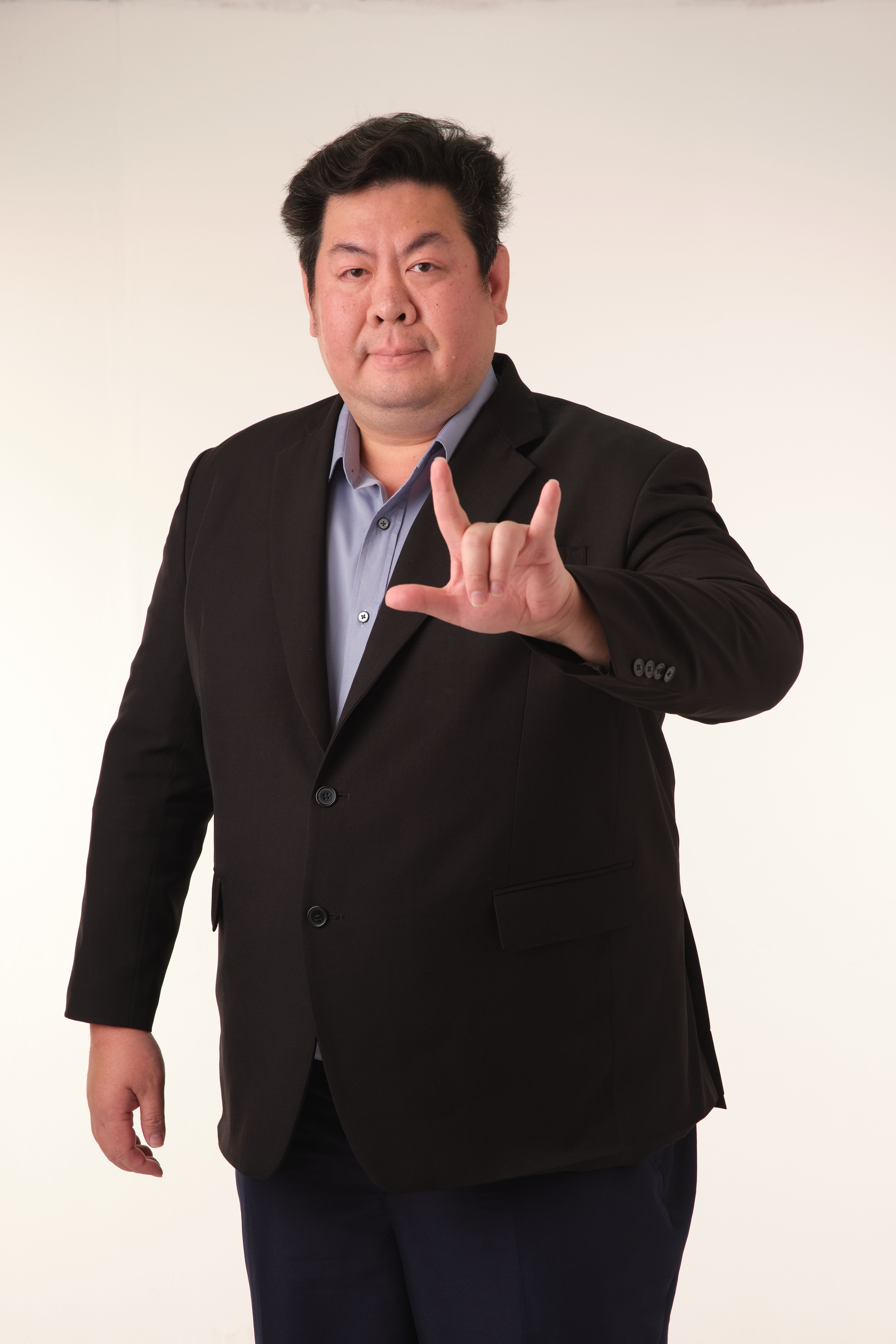 A confident adult man in a suit gestures 'I love you' in sign language.