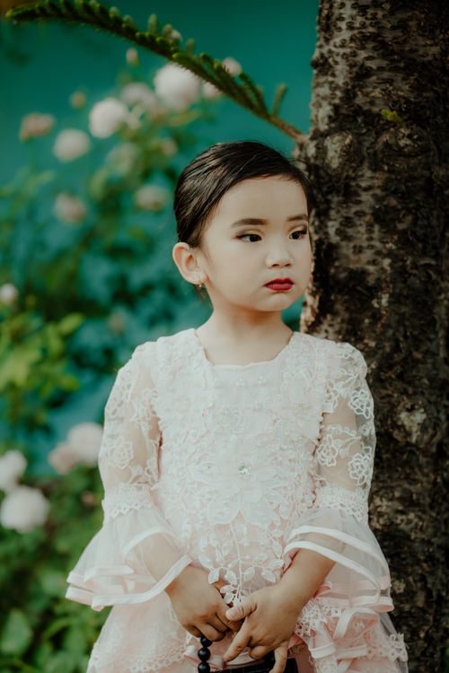 Free Beautiful child in a lace dress standing by a tree with flowers in the background. Stock Photo