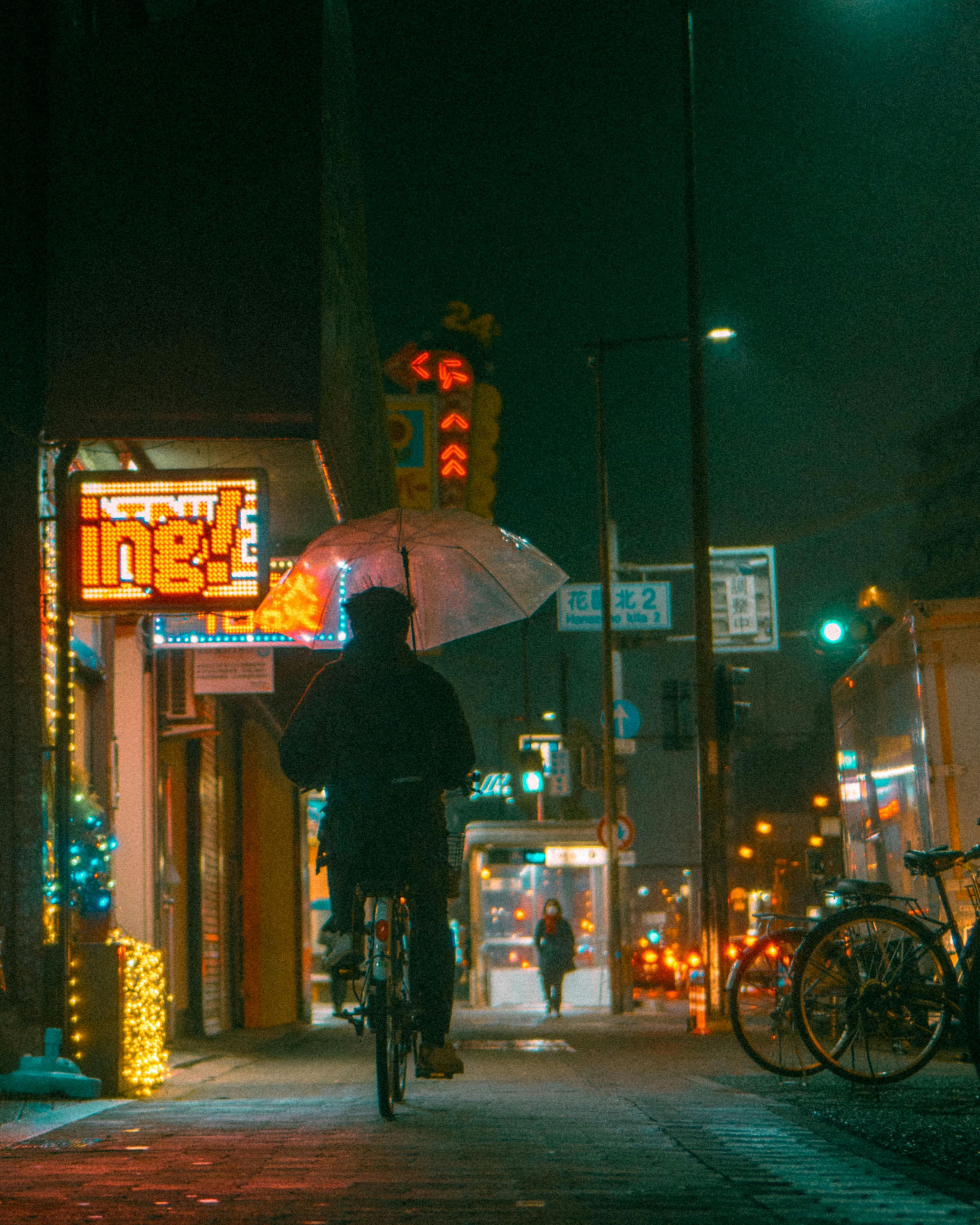 Atmospheric night photography of a cyclist and pedestrians in a lit Japanese street.