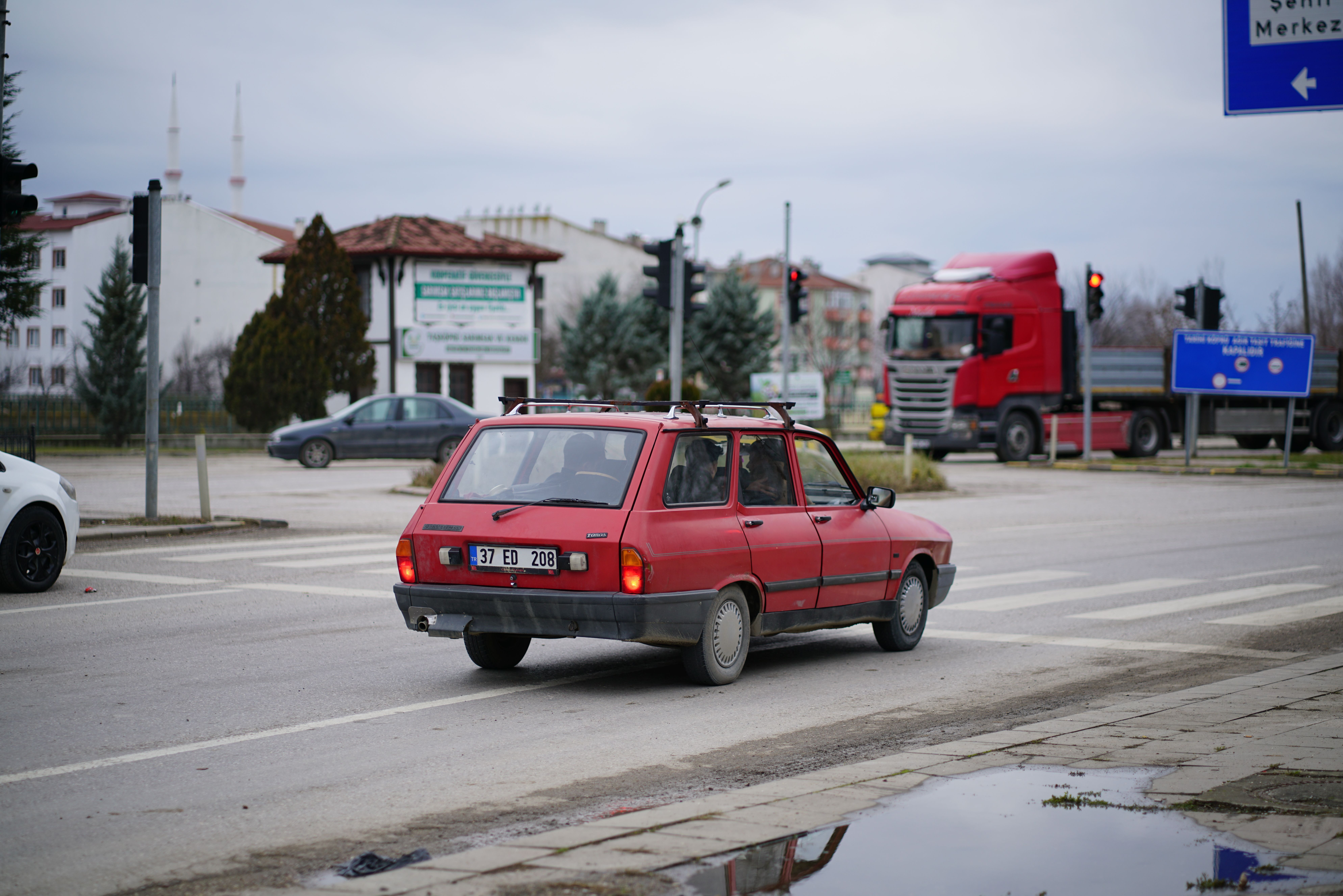 A vintage red car navigating an urban street with traffic signs and buildings in the background.