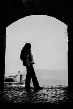 Black and white silhouette of a woman standing under an archway on a cobblestone path.