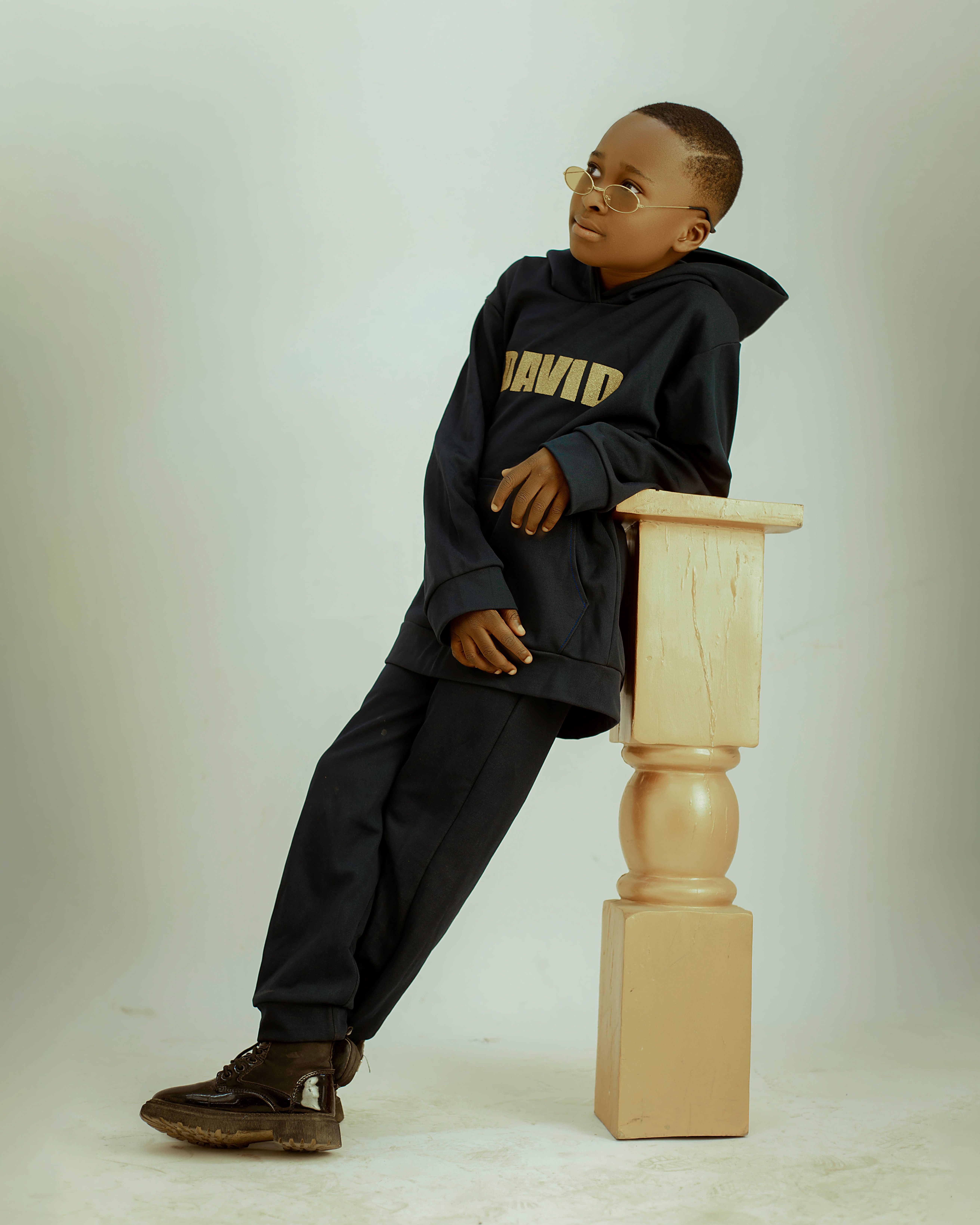 Fashionable child in glasses and navy outfit leaning casually on a wooden pedestal.