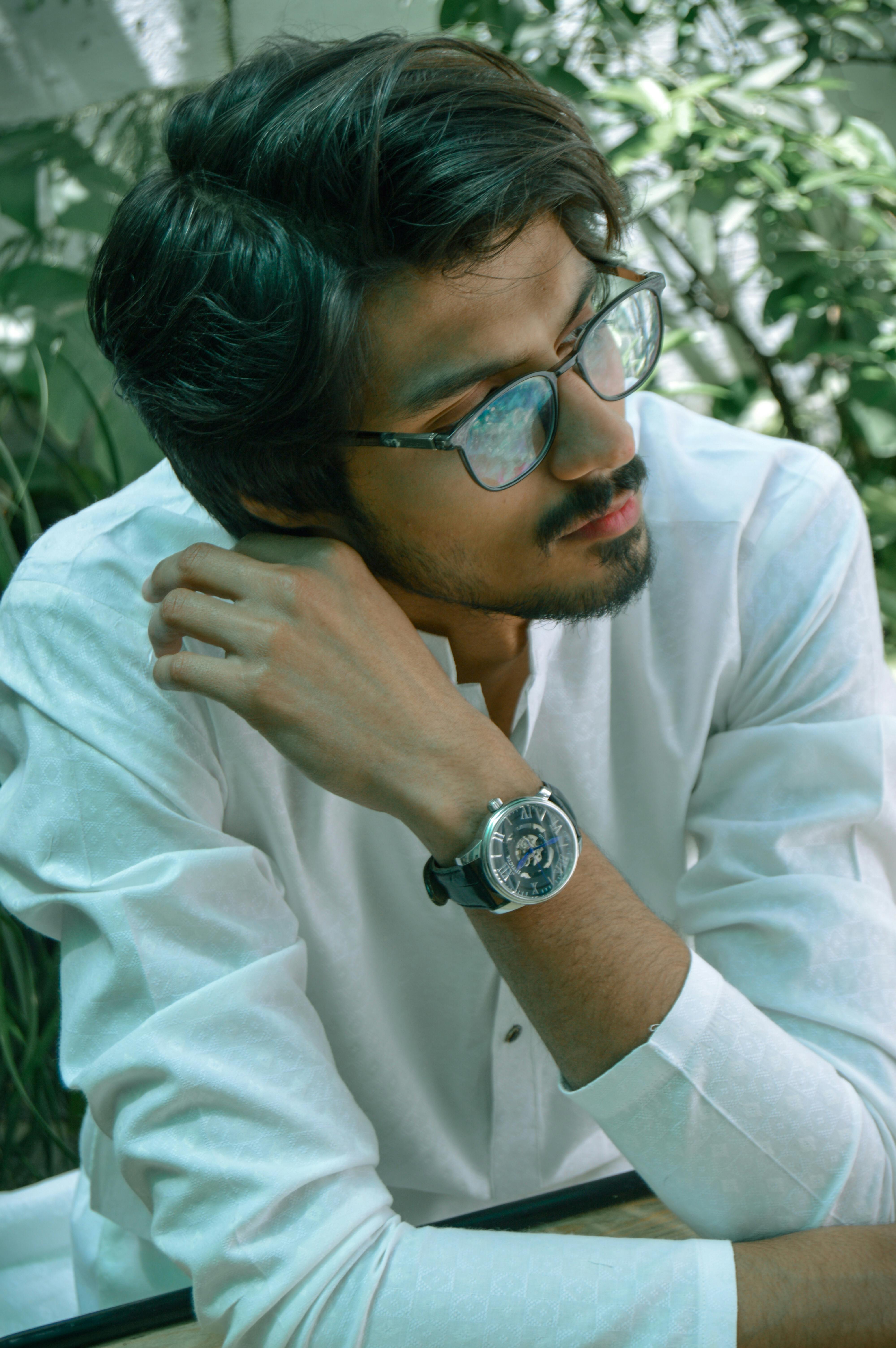 Man with glasses in a white shirt, sitting thoughtfully outdoors.