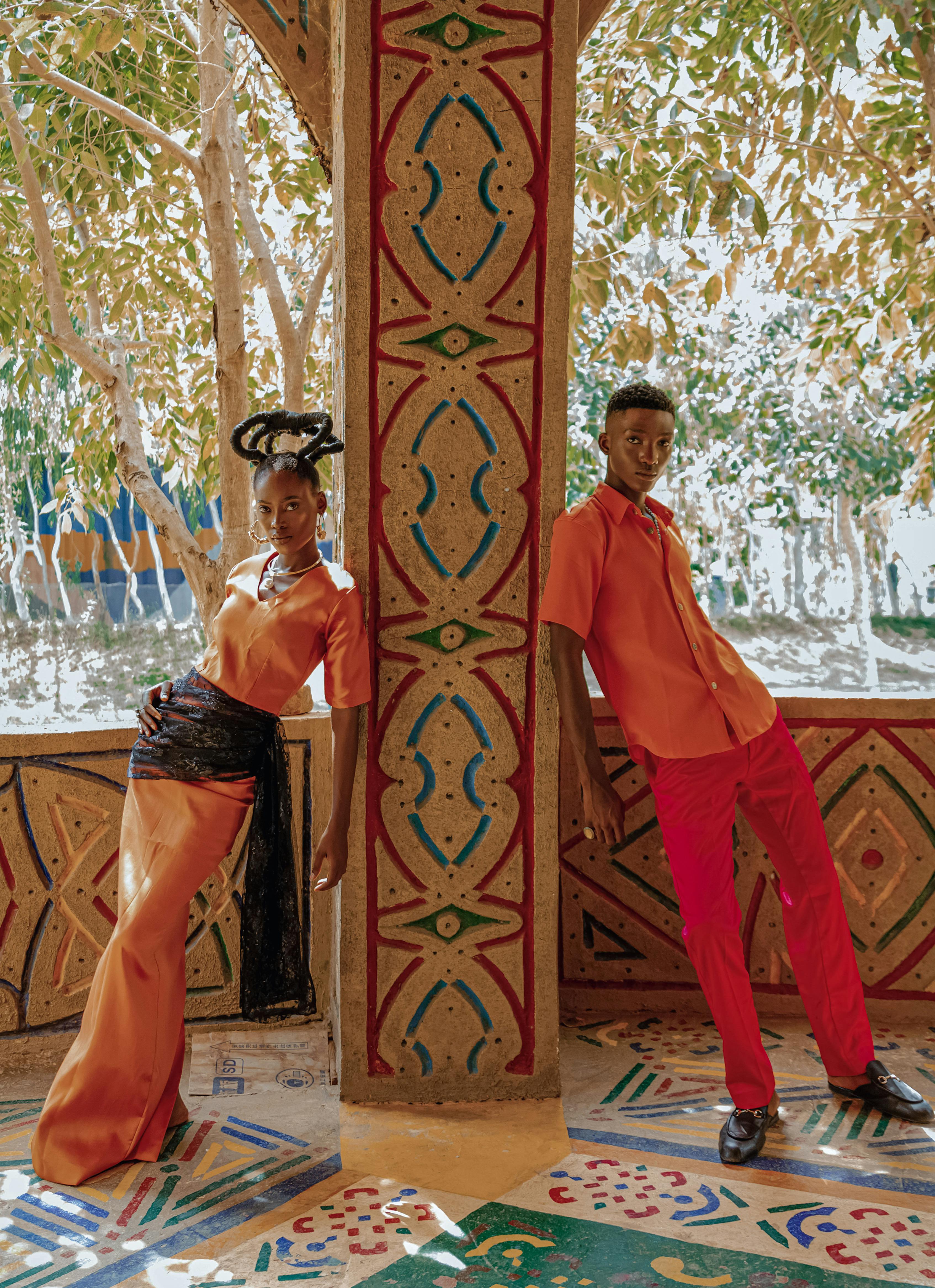 Colorful portrait of two adults in striking outfits against an ornate backdrop.