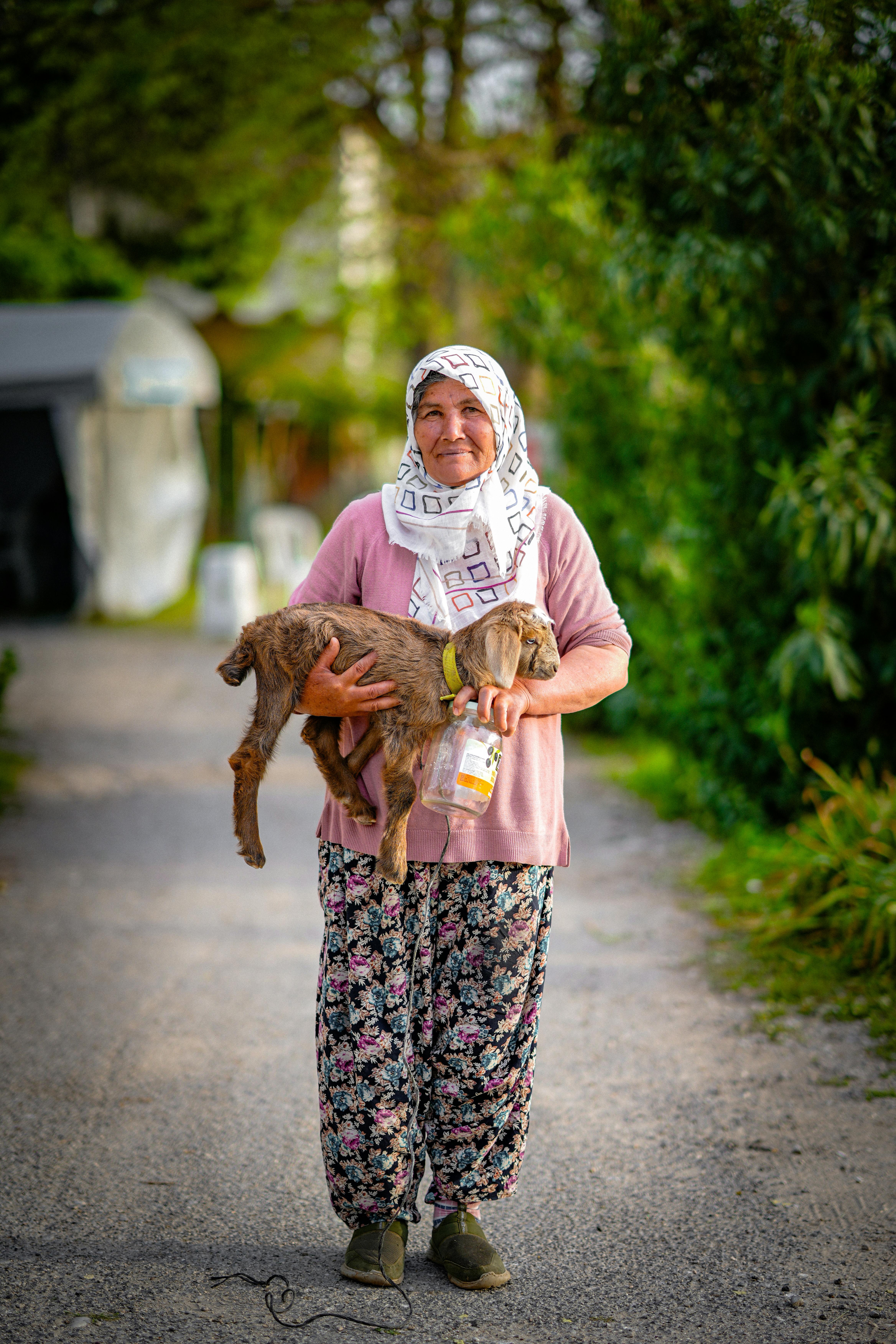 Elderly woman on a country road holding a small goat, smiling warmly.