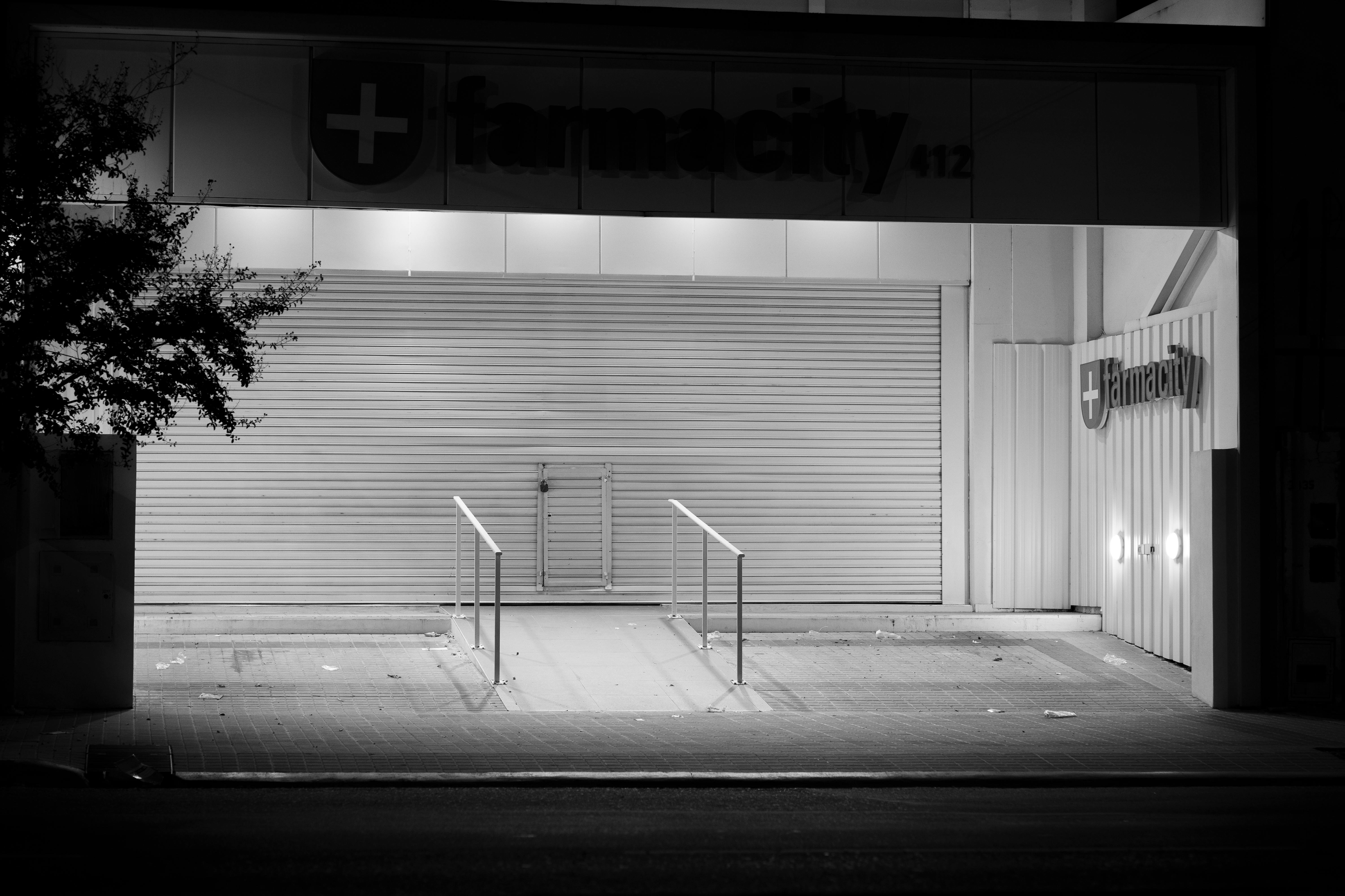 De franc Fotografia en blanc i negre d'una farmàcia tancada a Córdoba, Argentina, de nit, amb persianes metàl·liques i un arbre. Foto d'estoc