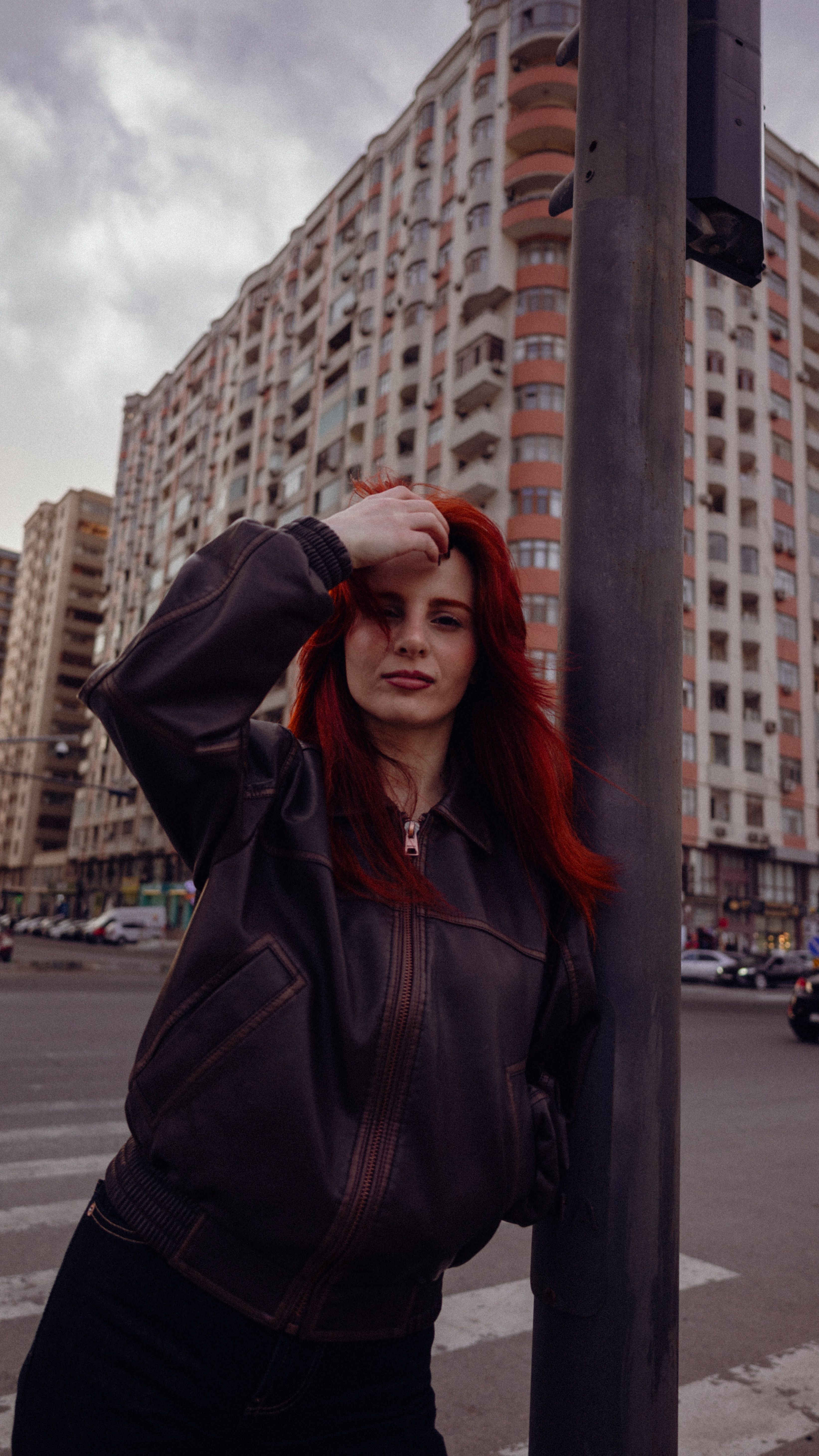 Stylish woman with red hair leans on pole in city street with tall buildings.