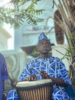 Elderly African man in traditional attire playing a djembe drum outdoors.